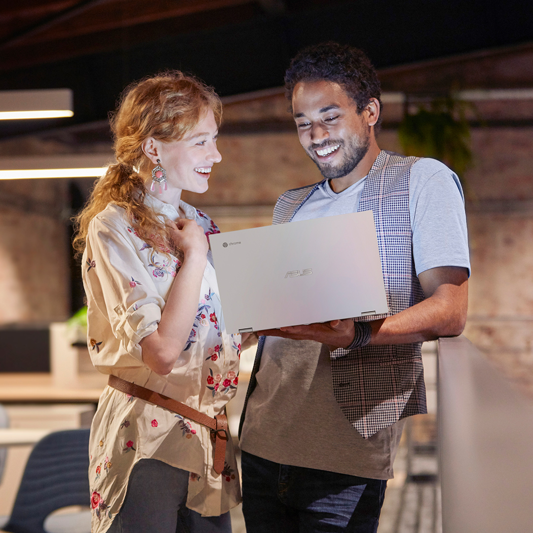 En kvinna och en man står bredvid varandra på ett modernt kontor. Mannen står till höger och håller en ASUS Chromebook framför sig.